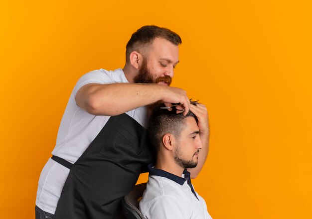 Professioneller bärtiger Friseurmann in der Schürze, die Haarschnitt mit Rasiermaschine zu einem jungen Mann macht, der über orange Wand steht