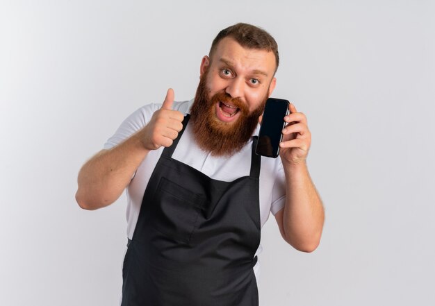 Professioneller bärtiger Friseurmann in der Schürze glücklich und aufgeregt, auf Handy zu sprechen, das Daumen oben zeigt über weißer Wand