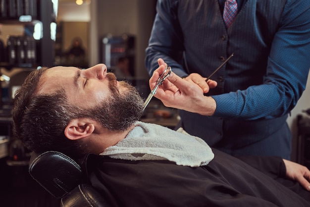 Professioneller friseur, der bart mit schere und kamm im friseursalon modelliert. nahaufnahmefoto.