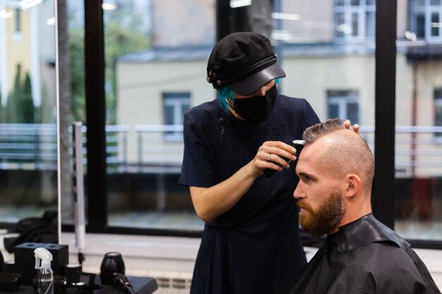 Professioneller Friseur, der eine schützende Gesichtsmaske trägt, machen Haarschnitt für europäischen bärtigen brutalen Mann im Schönheitssalon