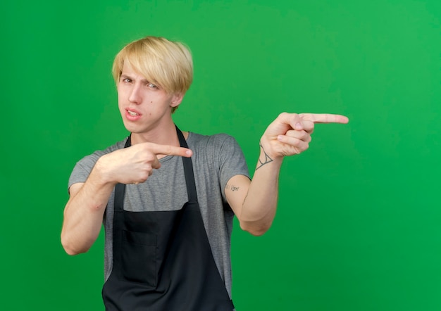 Kostenloses Foto professioneller friseur in der schürze, die mit zeigefingern zur seite mit ernstem gesicht zeigt