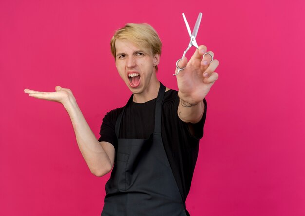 Professioneller Friseur in der Schürze hält eine Schere, die etwas mit dem Arm seiner Hand emotional und aufgeregt präsentiert