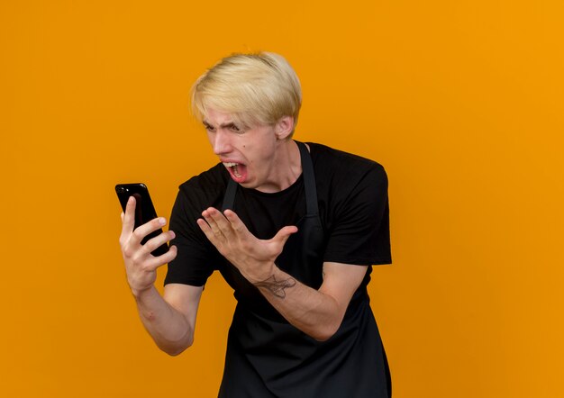 Professioneller Friseurmann in der Schürze, der Bildschirm seines Handys betrachtet, der mit aggressivem Ausdruck schreit, der frustriert ist, über orange Wand zu stehen