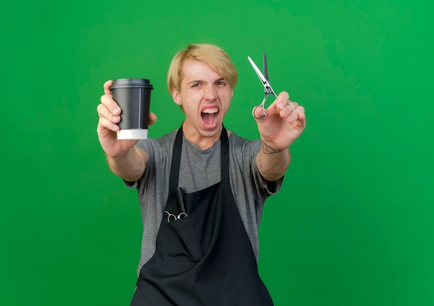 Professioneller Friseurmann in der Schürze, die Kaffeetasse und Schere schreit, die aufgeregt sind, wild zu gehen