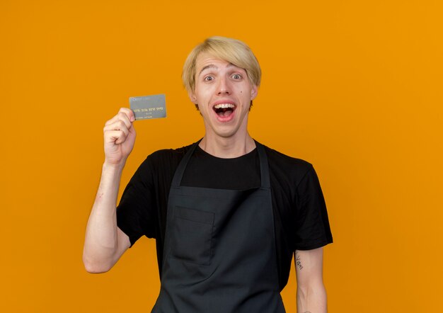 Kostenloses Foto professioneller friseurmann in der schürze, die kreditkarte hält, die mit glücklichem gesicht lächelt