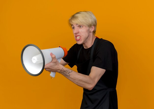 Professioneller Friseurmann in der Schürze, die Megaphon hält, das beiseite mit dem verärgerten Gesicht steht, das über orange Wand steht