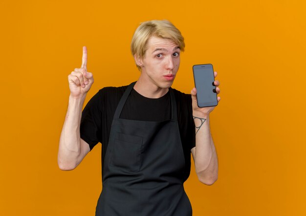 Professioneller Friseurmann in der Schürze, die Smartphone zeigt Zeigefinger, der zuversichtlich schaut, neue Idee zu haben, die über orange Wand steht