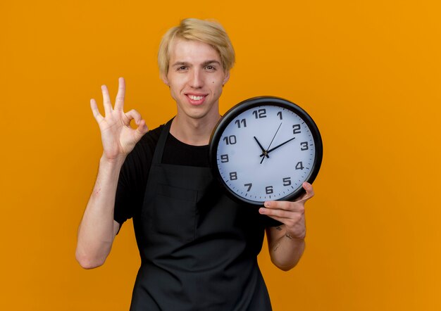 Professioneller Friseurmann in der Schürze, die Wanduhr hält, die nach vorne zeigt, das ok Zeichen steht, das über orange Wand steht