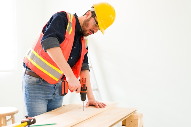 Professioneller Ingenieur und Auftragnehmer, der eine Holzplatte bohrt. Männlicher Tischler mit einem Schutzhelm, der am Bau einiger Möbel arbeitet