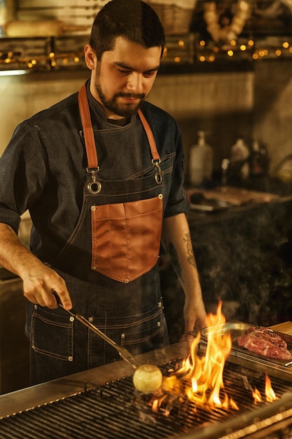 Kostenloses Foto professioneller koch kocht zwiebelgemüse und fleisch auf dem grill mit feuer und rauch schöner mann konzentrierte sich auf die zubereitung von speisen hintergrund der modernen restaurantküche