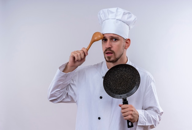 Professioneller männlicher Koch kocht in der weißen Uniform und im Kochhut, der den Kratzkopf der Bratpfanne mit dem Holzlöffel hält, der verwirrt steht, der über weißem Hintergrund steht