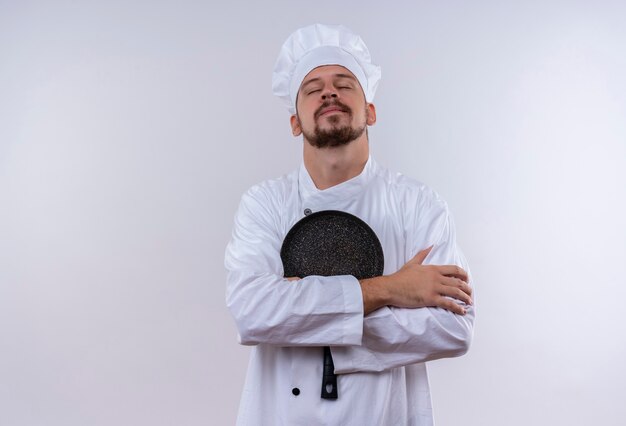 Professioneller männlicher Koch kocht in der weißen Uniform und im Kochhut, der die Bratpfanne hält, die selbstbewusst, selbstzufrieden steht und über weißem Hintergrund steht