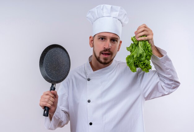 Professioneller männlicher Koch kocht in der weißen Uniform und im Kochhut, der die Bratpfanne und den lächelnden frischen Salat hält, der über weißem Hintergrund steht