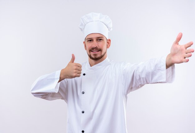 Professioneller männlicher Koch kocht in der weißen Uniform und im Kochhut, der die einladende Geste macht, die Daumen oben lächelnd freundlich stehend über weißem Hintergrund zeigt