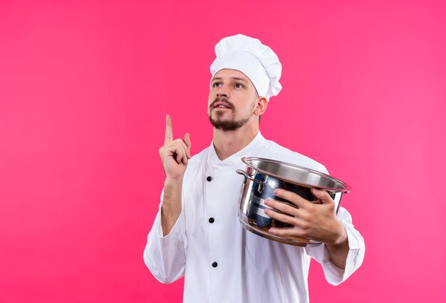 Professioneller männlicher Koch kocht in der weißen Uniform und im Kochhut, der eine leere Pfanne hält, die oben zeigenden Finger nach oben hat und neue Idee hat, die über rosa Hintergrund steht