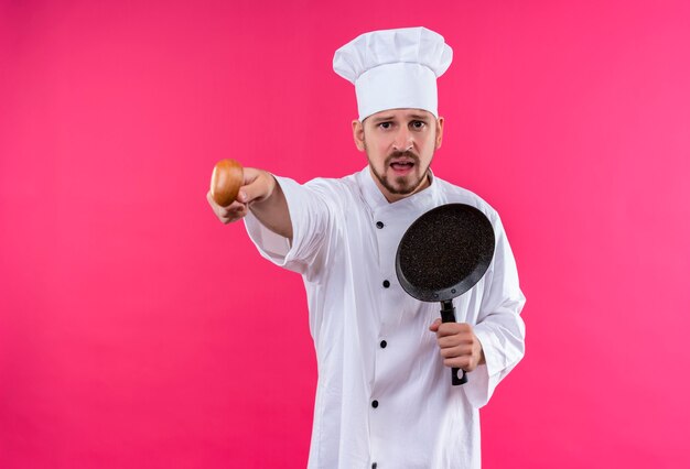Professioneller männlicher Koch kocht in der weißen Uniform und im Kochhut, der eine Pfanne hält, die auf etwas mit einem emotionalen und besorgten Holzlöffel zeigt, der über rosa Hintergrund steht