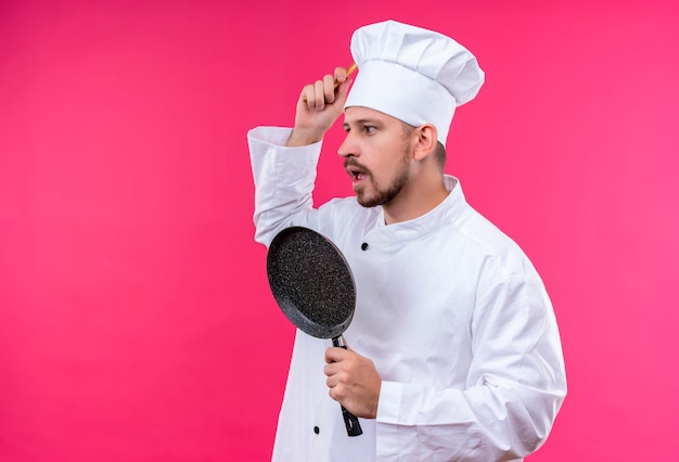 Professioneller männlicher Koch kocht in der weißen Uniform und im Kochhut, der eine Pfanne hält, die verwirrt und sehr ängstlich steht, die über rosa Hintergrund stehen