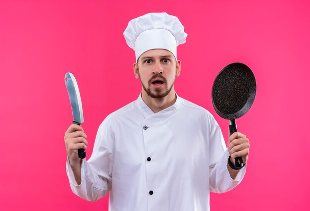 Professioneller männlicher Koch kocht in der weißen Uniform und im Kochhut, der eine Pfanne und ein Messer hält, die Kamera betrachten, die über rosa Hintergrund steht