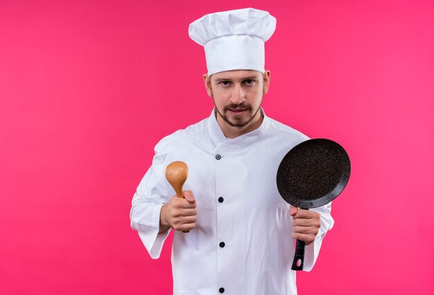 Professioneller männlicher Koch kocht in der weißen Uniform und im Kochhut, der eine Pfanne und einen Holzlöffel hält, die Kamera mit ernstem Gesicht betrachten, das über rosa Hintergrund steht