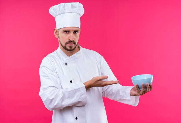 Professioneller männlicher Koch kocht in der weißen Uniform und im Kochhut, der eine Schüssel mit Arm seiner Hand darstellt, die über rosa Hintergrund steht