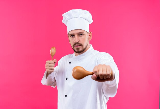 Professioneller männlicher Koch kocht in der weißen Uniform und im Kochhut, der hölzerne Löffel hält, die Kamera mit ernstem Gesicht betrachten, das über rosa Hintergrund steht