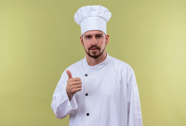 Kostenloses Foto professioneller männlicher koch kocht in der weißen uniform und im kochhut, der kamera betrachtet, die daumen oben steht über grünem hintergrund