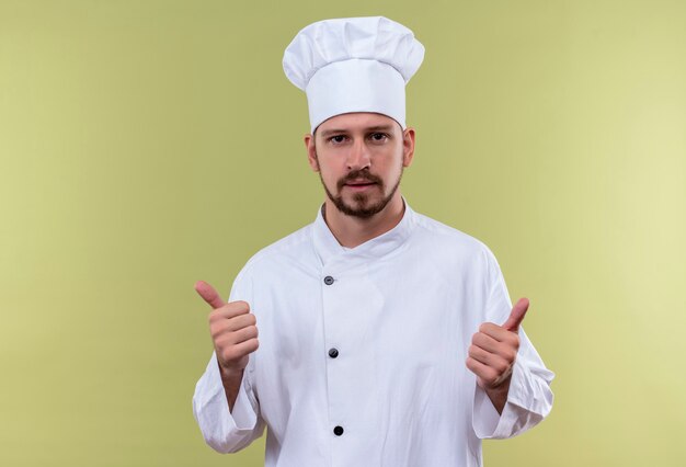 Professioneller männlicher Koch kocht in der weißen Uniform und im Kochhut, der Kamera betrachtet, die Daumen oben steht über grünem Hintergrund