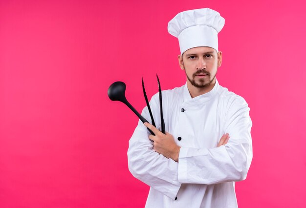 Professioneller männlicher Koch kocht in der weißen Uniform und im Kochhut, der Schöpflöffel hält Kamera mit ernstem Gesicht, das über rosa Hintergrund steht