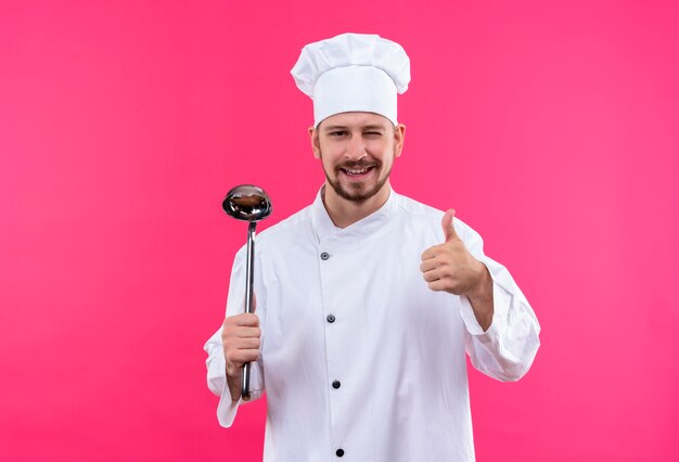 Professioneller männlicher Koch kocht in der weißen Uniform und im Kochhut, der Schöpflöffel hält lächelnd und zwinkernd zeigt Daumen hoch stehend über rosa Hintergrund