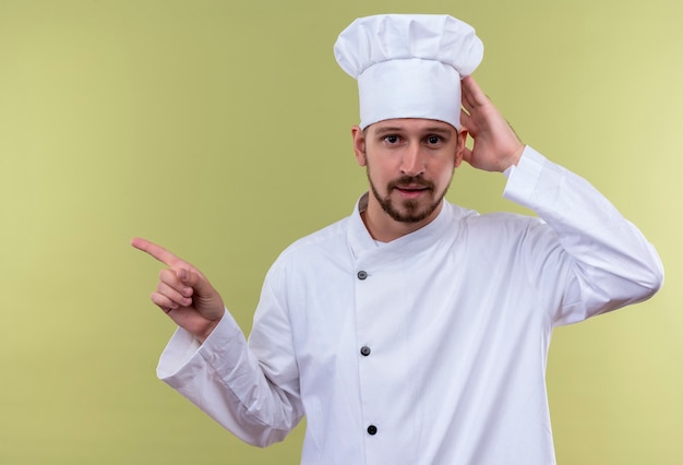 Professioneller männlicher Koch kocht in der weißen Uniform und im Kochhut, der seinen Hut berührt, der mit Zeigefinger zur Seite zeigt, mit dem selbstbewussten Lächeln auf Gesicht, das über grünem Hintergrund steht