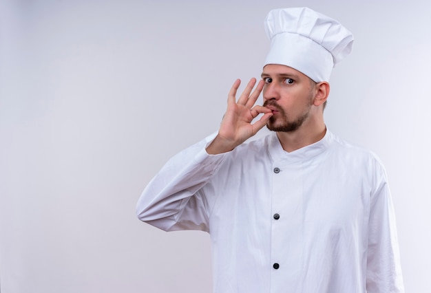 Professioneller männlicher Koch kocht in der weißen Uniform und im Kochhut, der Zeichen für köstliches Stehen über weißem Hintergrund zeigt