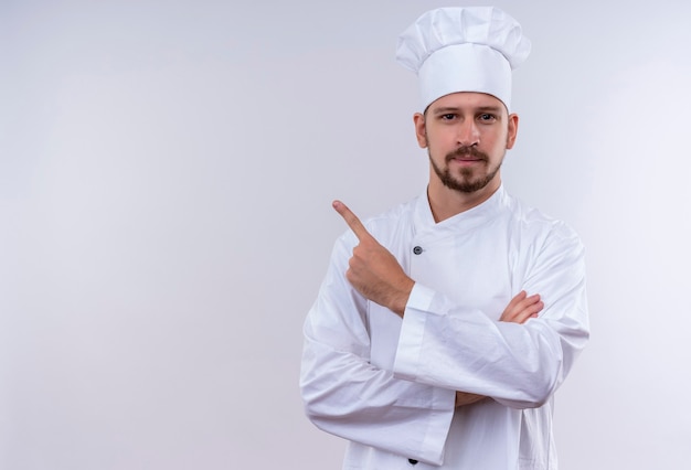 Kostenloses Foto professioneller männlicher koch kocht in der weißen uniform und im kochhut, der zur seite mit zeigefinger zeigt, der zuversichtlich steht, über weißem hintergrund zu stehen