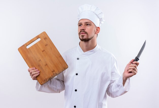 Professioneller männlicher Kochkoch in der weißen Uniform und im Kochhut, der das hölzerne Schneidebrett und das Messer hält, die beiseite mit nachdenklichem Ausdruck auf Gesicht stehen, das über weißem Hintergrund steht