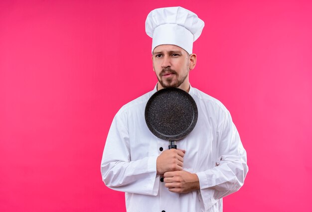 Professioneller männlicher Kochkoch in der weißen Uniform und im Kochhut, der eine Pfanne hält, die beiseite mit skeptischem Ausdruck auf Gesicht steht, das über rosa Hintergrund steht