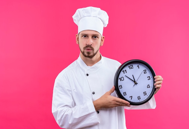Professioneller männlicher Kochkoch in der weißen Uniform und im Kochhut, der eine Uhr hält, die Kamera mit sicherem Ausdruck betrachtet, der über rosa Hintergrund steht