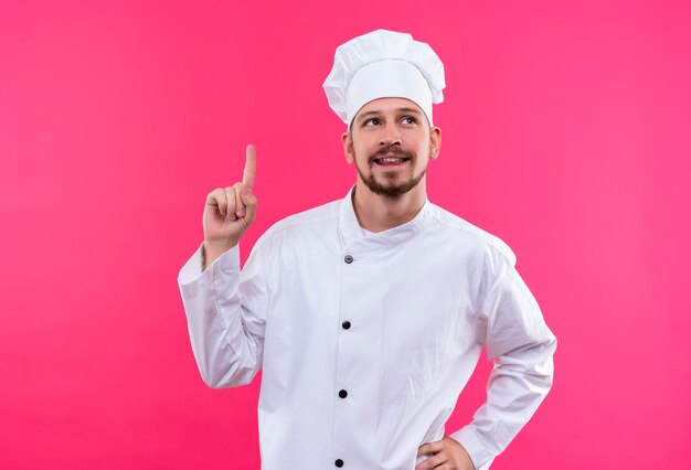 Professioneller männlicher Kochkoch in der weißen Uniform und im Kochhut, der oben zeigt und mit dem Finger zeigt, erinnert sich nicht daran, wichtige Sache zu vergessen, die über rosa Hintergrund steht