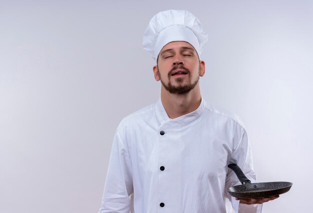 Professioneller männlicher Kochkoch in der weißen Uniform und im Kochhut stehend mit geschlossenen Augen, die Bratpfanne über weißem Hintergrund halten