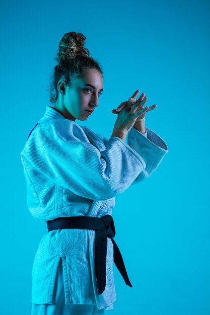 Professioneller weiblicher Judoist im weißen Judo-Kimono, der lokalisiert auf blauem neoniertem Studiohintergrund praktiziert und trainiert.