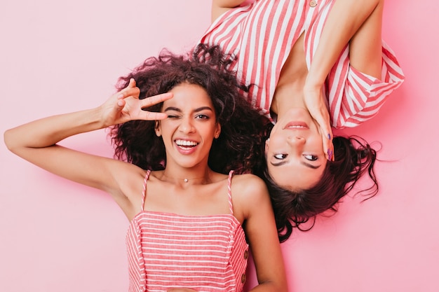 Professionelles Foto von jungen und attraktiven Mädchen mit schönem Make-up und dunklem lockigem Haar. Frauen liegen auf dem Boden, spielen herum und zeigen Friedenszeichen.