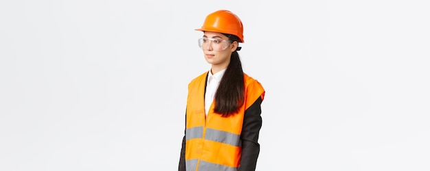 Profil einer seriösen asiatischen Geschäftsfrau, die den Chefingenieur des Baubereichs inspiziert und nach links schaut und einen Schutzhelm und reflektierende Kleidung auf weißem Hintergrund trägt