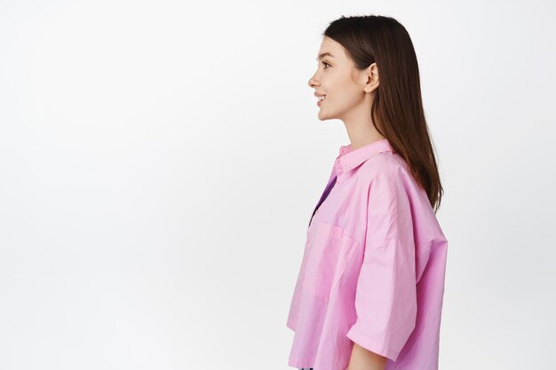 Profilaufnahme eines brünetten lächelnden Mädchens in rosafarbenem Hemd, das nach links schaut, mit einem glücklichen, entspannten Gesichtsausdruck, der auf weißem Hintergrund steht. Kopieren Sie Platz