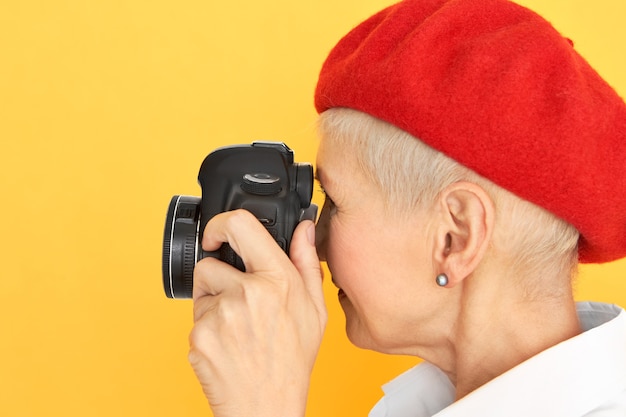 Profilporträt des kreativen stilvollen kurzhaarigen Frauenfotografen mittleren Alters in der roten Haube, die gegen gelben Hintergrund mit professioneller Digitalkamera in ihren Händen aufwirft. Kunstfotografie