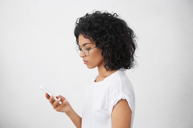 Profilporträt einer gut aussehenden attraktiven schwarzen Mischlingsfrau in einer runden Brille, die ein weißes Smartphone in der Hand hält und es mit ernstem Gesichtsausdruck verwendet und nach wichtigen Informationen sucht