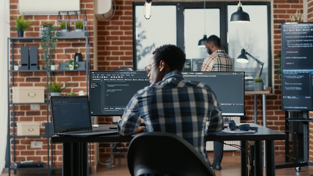 Programmierer-Codierung auf einem Laptop, der sich an einen Schreibtisch mit Computerbildschirmen setzt, die Code in einer Software-Agentur parsen. Softwareentwickler, der Algorithmen kompiliert, während Cloud-Programmierer im Hintergrund arbeiten.