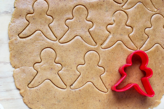 Prozess der Behandlung von Lebkuchenmannplätzchen, benutzen roten Lebkuchenmann-Formausschnitt-Lebkuchenteig auf Backpapier um bunte Ausstechformen auf weißem Holztisch. Ansicht von oben