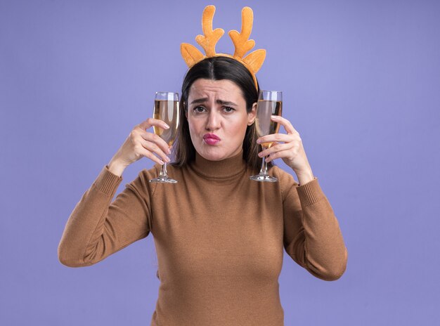 Pursing Lippen junges schönes Mädchen, das braunen Pullover mit Weihnachtshaarbügel hält, der zwei Glas Champagner um Ohren lokalisiert auf blauem Hintergrund hält