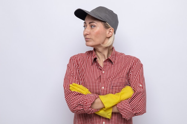 Putzfrau in kariertem hemd und mütze mit gummihandschuhen, die mit ernstem gesicht mit verschränkten armen auf weißem hintergrund zur seite schaut