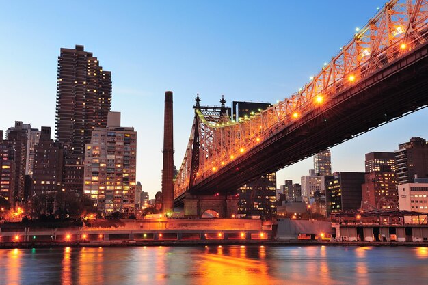 Queensboro Bridge über New York City East River bei Sonnenuntergang mit Reflexionen des Flusses und Skyline von Midtown Manhattan beleuchtet.