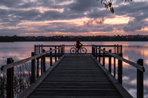 Radfahrer, der auf einem hölzernen Dock auf dem Wasser unter einem bewölkten Himmel während des Sonnenuntergangs am Abend steht