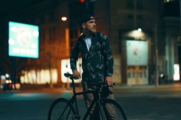 Radfahrer, die neben seinem klassischen Fahrrad auf der Straße stehen, während sie die Nachtstadt beobachten